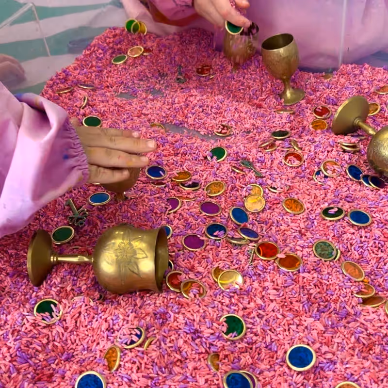 Hands digging through a tray of pink sensory rice and treasure