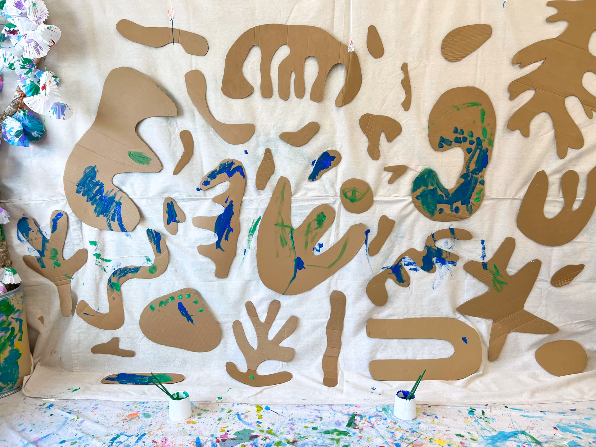 A wall of large cardboard cutout creatures in painterly shapes, with smudges of blue and green paint, ready for an excursion painting session.