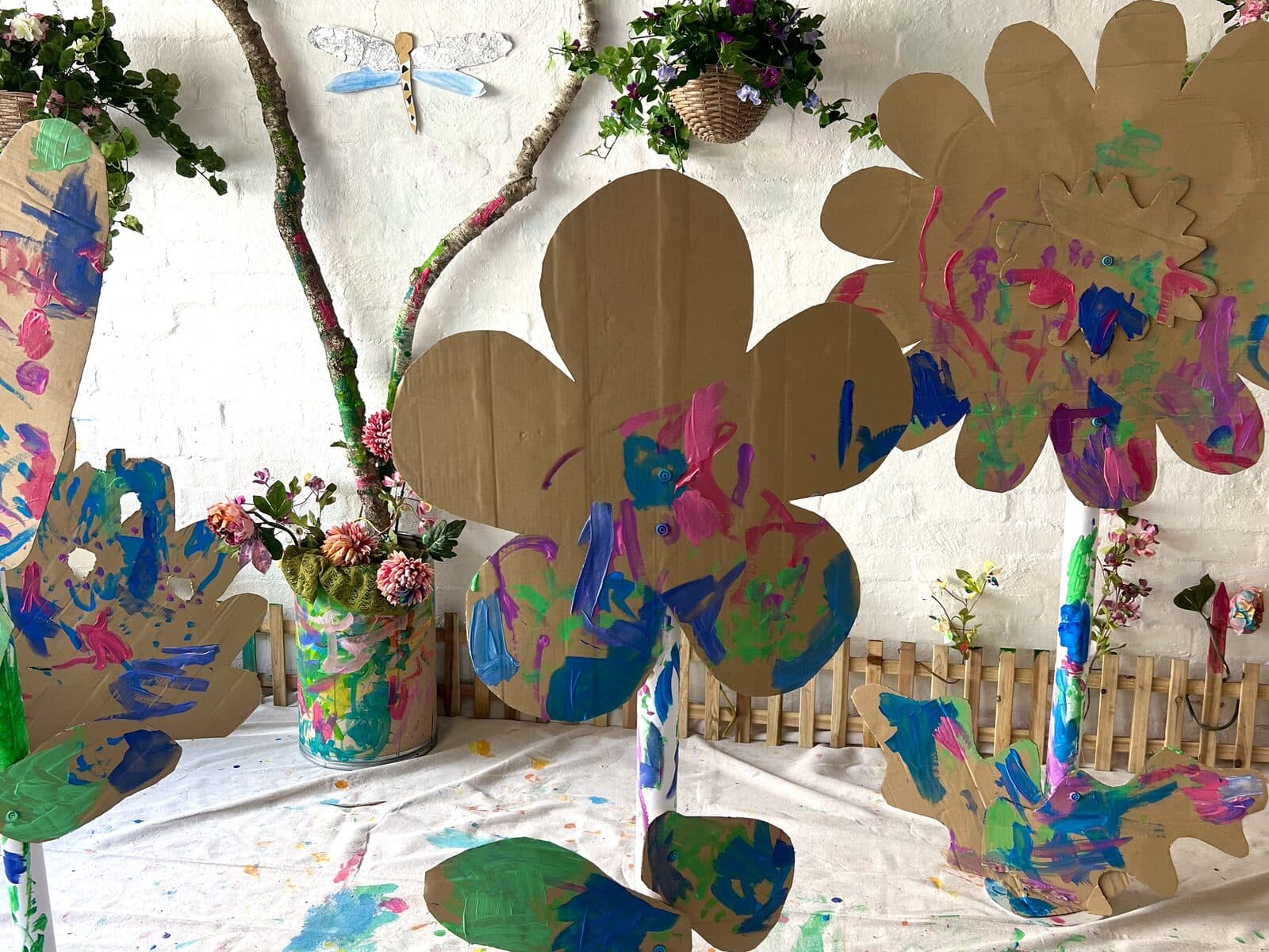 Giant cardboard flowers painted in pink, blue and green, standing on a paint-splattered drop sheet against a white brick wall.