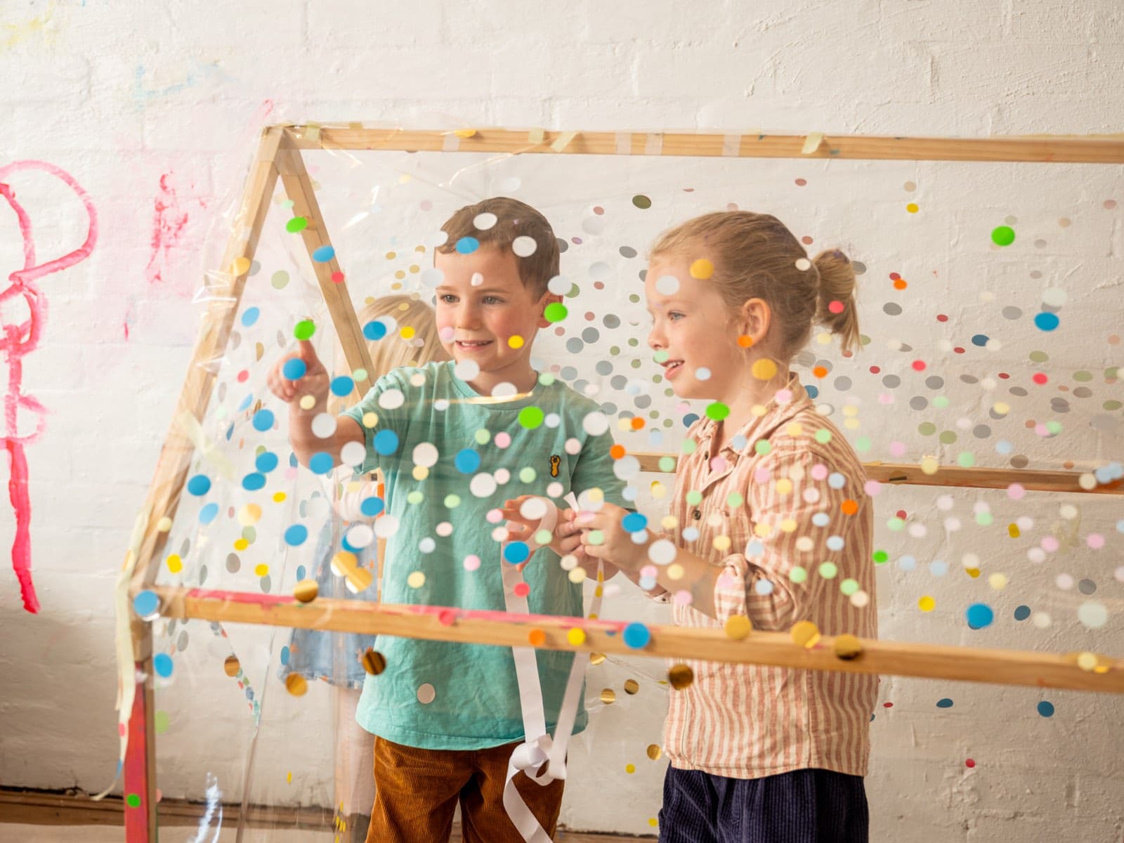 Two children threading ribbon through a cubby-house frame wrapped in clear film covered with colourful confetti dots, at the Smudge studio